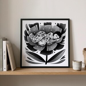 May include: Black and white art print of a person sleeping inside a large flower. The person is covered in floral tattoos. The artwork is framed in a black frame and displayed on a wooden shelf with books and a mug.
