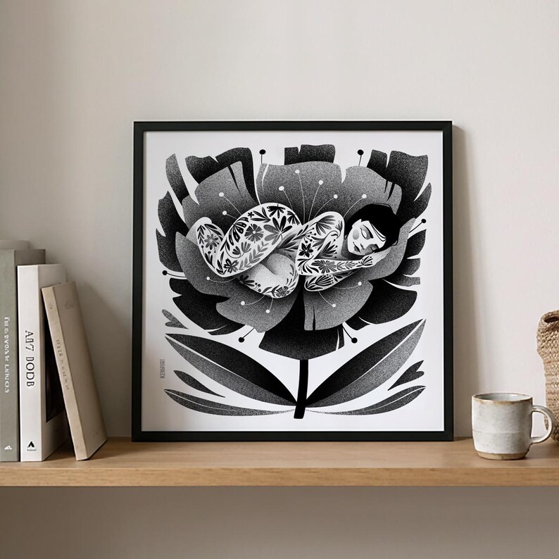 May include: Black and white art print of a person sleeping inside a large flower. The person is covered in floral tattoos. The artwork is framed in a black frame and displayed on a wooden shelf with books and a mug.