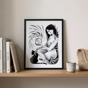 May include: Black and white art print in a black frame. The artwork depicts a seated figure holding a glowing orb, with a black cat at their side. Botanical elements surround the figure. The print is displayed on a wooden shelf with books and a mug.