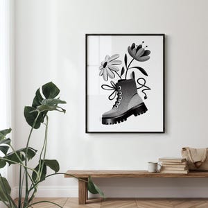May include: Black and white framed art print featuring a combat boot with flowers. The boot is gray with black accents and laces, and the flowers are stylized. The artwork is displayed on a white wall.