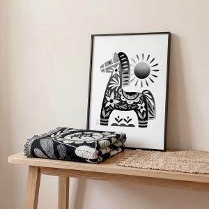 以下が含まれることがあります： 花柄と太陽が描かれた装飾的な馬の白黒フレームプリント。折りたたまれた白黒の花柄のスローブランケットが木製の表面に置かれ、織られたマットの隣にあります。