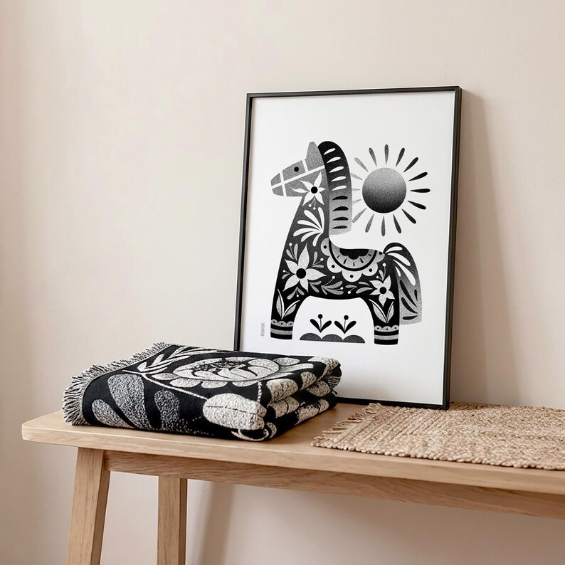 以下が含まれることがあります： 花柄と太陽が描かれた装飾的な馬の白黒フレームプリント。折りたたまれた白黒の花柄のスローブランケットが木製の表面に置かれ、織られたマットの隣にあります。