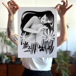 以下が含まれることがあります： 長い髪の座っている人物が、様式化された花と葉に囲まれた白黒のアートプリント。アートワークは柔らかく点描のテクスチャで、夢のような雰囲気を作り出しています。人物は瞑想的なポーズをとっています。