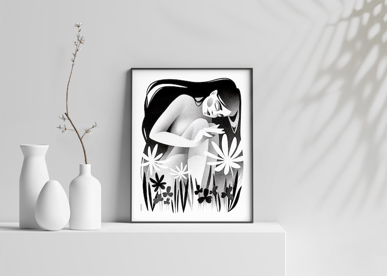 以下が含まれることがあります： 長い髪の女性が様式化された花の中に座っている白黒のアートプリント。アートワークは額装され、白い表面に展示されており、近くには装飾用の白い花瓶と花の小枝があります。