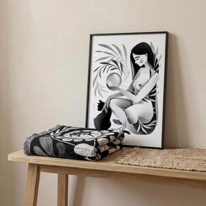 May include: A black and white framed art print of a woman holding a sphere, with a black cat, and a folded black and white floral throw blanket. The art print and blanket are on a light wood table.