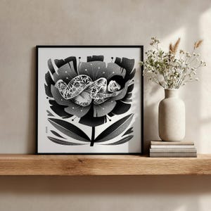 May include: Black and white art print of a person sleeping inside a large flower. The person has floral tattoos. The flower is detailed with dark and light gray shading. The print is in a black frame and sits on a wooden shelf.