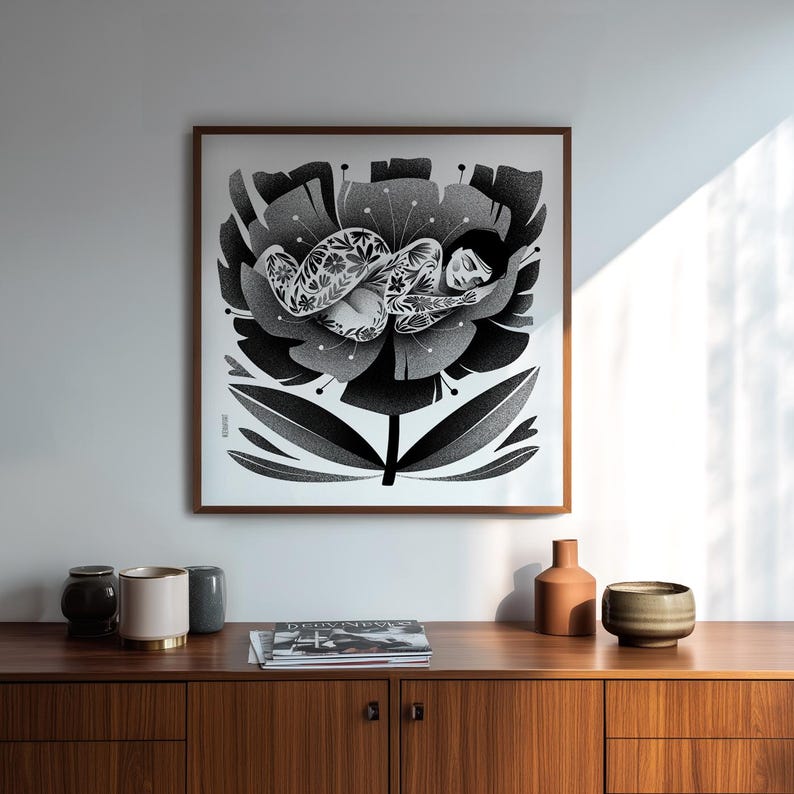 May include: Black and white art print of a person sleeping inside a large flower. The person is adorned with floral tattoos. The artwork is framed in a brown wooden frame and hangs on a white wall. The print is displayed above a wooden cabinet.