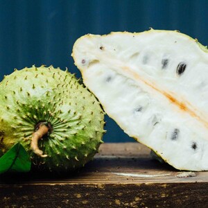 Peut inclure: Un corossol entier vert &agrave; l'ext&eacute;rieur &eacute;pineux avec une feuille verte &agrave; c&ocirc;t&eacute; d'un corossol coup&eacute; en deux r&eacute;v&eacute;lant une chair blanche fibreuse et des graines brun fonc&eacute;. Le fruit est pos&eacute; sur une surface en bois sombre devant un fond bleu.