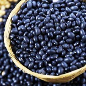 Puede incluir: Una cesta tejida llena de peque&ntilde;os frijoles negros de forma ovalada. Los frijoles tienen una superficie oscura y lisa, y est&aacute;n densamente empaquetados en el recipiente. Se ven frijoles negros adicionales y una porci&oacute;n de frijoles beige.