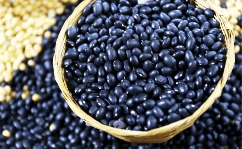 Puede incluir: Una cesta tejida llena de peque&ntilde;os frijoles negros de forma ovalada. Los frijoles tienen una superficie oscura y lisa, y est&aacute;n densamente empaquetados en el recipiente. Se ven frijoles negros adicionales y una porci&oacute;n de frijoles beige.