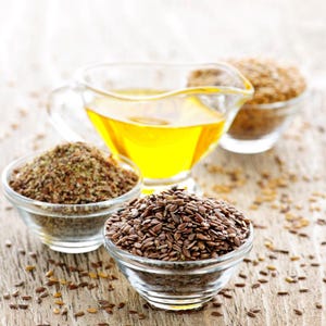 Pu&ograve; includere: Una caraffa in vetro trasparente riempita di olio di semi di lino giallo dorato posizionata dietro due piccole ciotole di vetro, una contenente semi di lino marroni interi e l'altra semi di lino macinati, con semi sparsi sulla superficie.