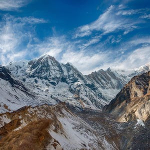 May include: A vast mountain range featuring jagged, snow-covered peaks under a bright blue sky with wispy white clouds. The slopes display a mix of white snow, exposed dark grey rock, and patches of dry, golden-brown vegetation in the foreground.
