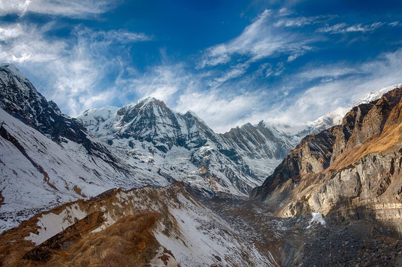 May include: A vast mountain range featuring jagged, snow-covered peaks under a bright blue sky with wispy white clouds. The slopes display a mix of white snow, exposed dark grey rock, and patches of dry, golden-brown vegetation in the foreground.