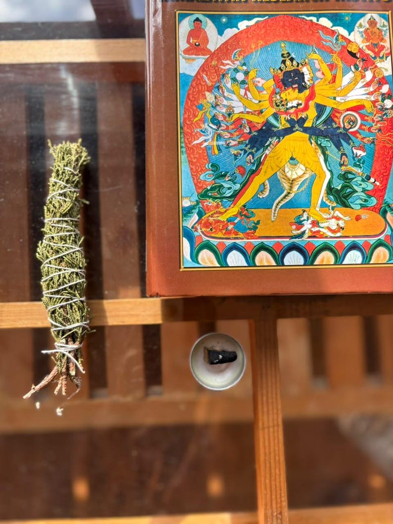 May include: A bundle of dried green herbs tied with white string, a small circular metal incense holder with a dark cone, and a rectangular book featuring a colorful illustration of a multi-armed deity in yellow and blue tones.