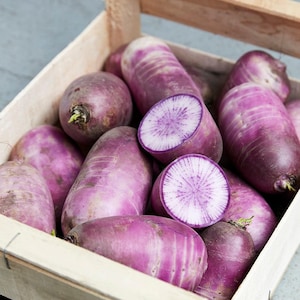 Puede incluir: Una colecci&oacute;n de r&aacute;banos daikon morados empacados dentro de una caja de madera clara. Varios r&aacute;banos est&aacute;n enteros con una piel morada vibrante y suave, mientras que dos est&aacute;n cortados transversalmente revelando un interior blanco con patrones radiales morados.
