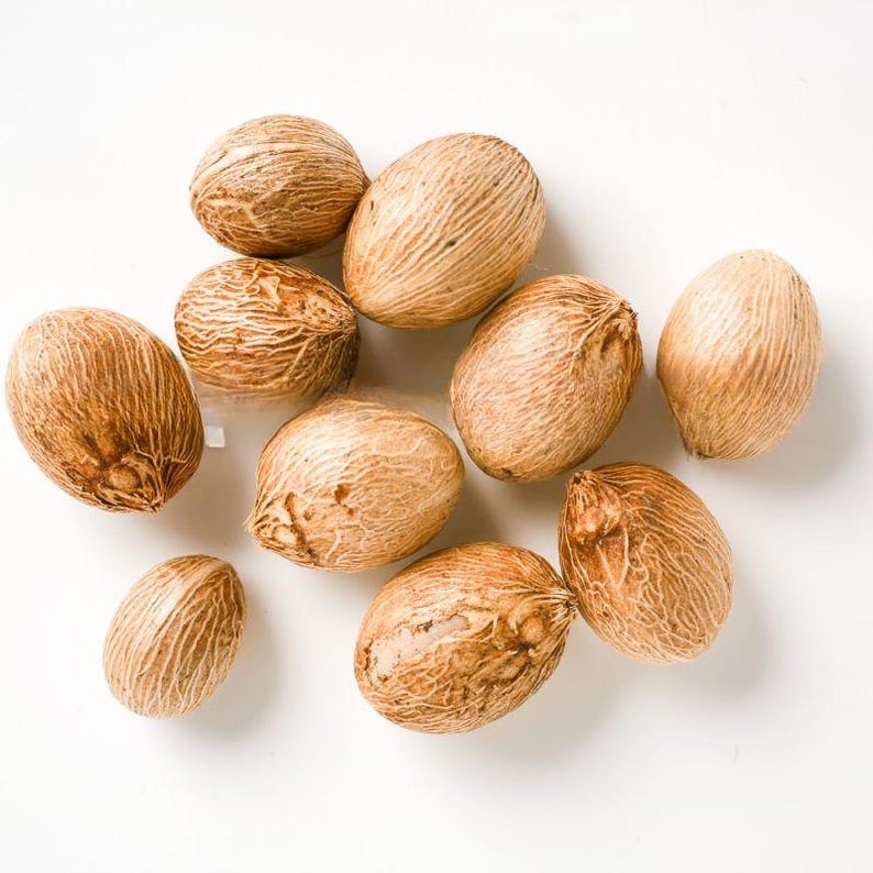 Puede incluir: Una colecci&oacute;n de diez nueces moscadas enteras dispersas por el encuadre. Cada semilla tiene forma ovalada con un exterior le&ntilde;oso de color marr&oacute;n claro, que presenta una textura fibrosa de l&iacute;neas finas y una superficie de aspecto mate y ligeramente rugoso.