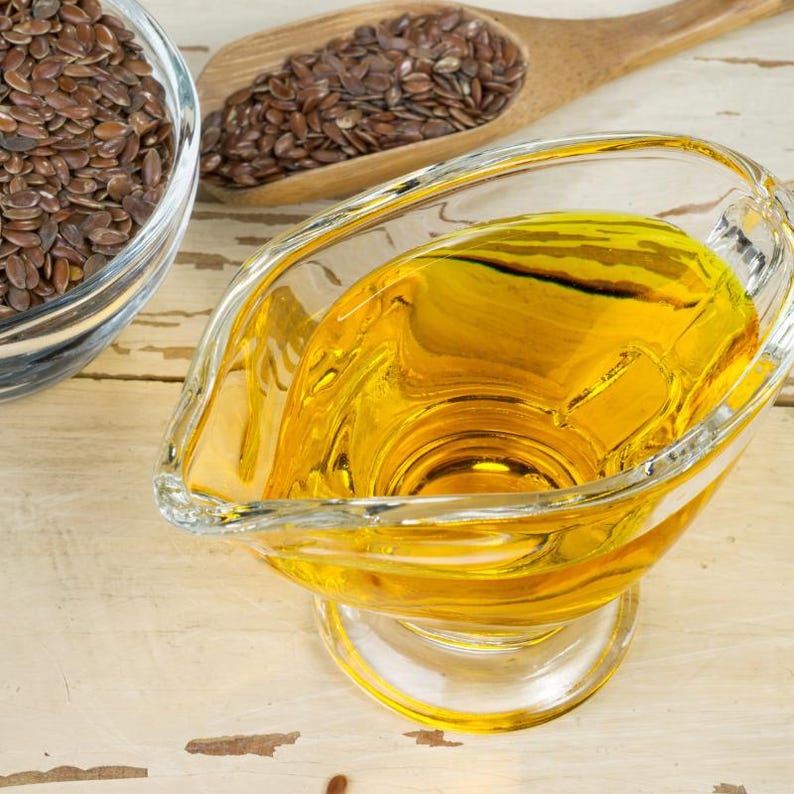 Pu&ograve; includere: Una salsiera in vetro trasparente riempita di olio di semi di lino giallo dorato, posizionata accanto a una ciotola di vetro contenente semi di lino marroni e un cucchiaio di legno pieno di semi, su una superficie in legno chiaro.