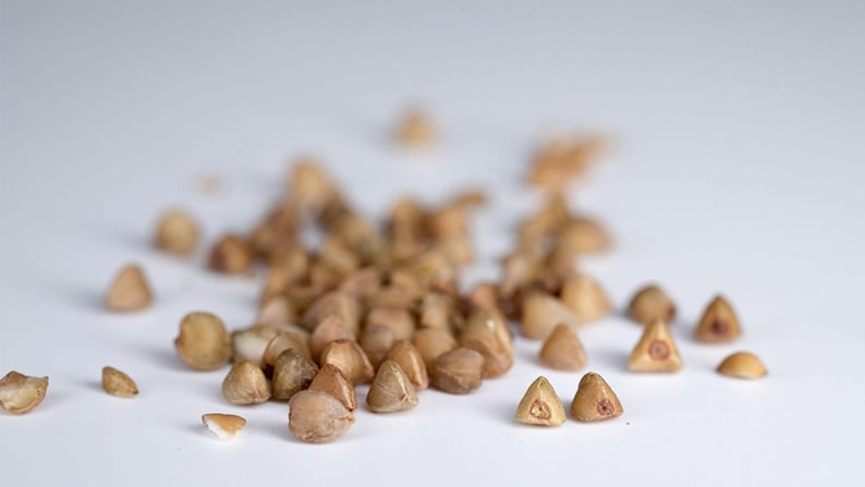 May include: A scattered pile of raw buckwheat groats, each featuring a distinct triangular shape with a light brown, tan, and beige color palette. The individual grains have a hard, hull-less texture and a small, darker center point on their flat faces.
