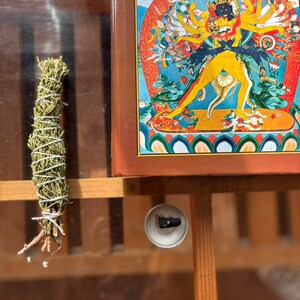 May include: A bundle of dried green herbs tied with white string, a small circular metal incense holder with a dark cone, and a rectangular book featuring a colorful illustration of a multi-armed deity in yellow and blue tones.