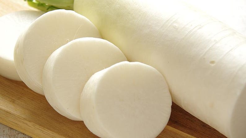 Op de afbeelding: Een lange, cilindervormige witte daikon-radijs met een gladde, stevige schil. De groente is gedeeltelijk in verschillende dikke, gelijkmatige, ronde schijven gesneden, wat een solide, ondoorzichtige witte binnenkant laat zien.