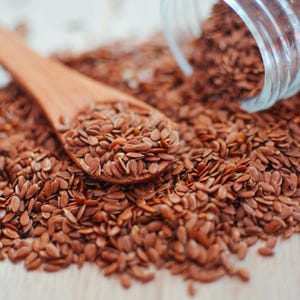 Pu&ograve; includere: Un cucchiaio di legno riempito con piccoli semi di lino marroni di forma ovale che poggia su un mucchio di semi sfusi. Un barattolo di vetro trasparente &egrave; rovesciato, versando altri semi di lino marroni sulla superficie, creando una trama naturale.
