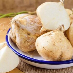 Pu&ograve; includere: Una ciotola bianca con bordo blu contenente diverse radici di jicama intere e una radice di jicama tagliata. Le radici hanno una buccia sottile color marrone chiaro e una forma arrotondata. Due fette triangolari di jicama bianca sono poste accanto.