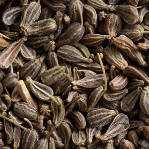 K&ouml;nnte beinhalten: Eine Ansammlung getrockneter Anissamen, die jeweils eine ausgepr&auml;gte gerippte, tropfenf&ouml;rmige Struktur mit einem br&auml;unlich-grauen Farbton aufweisen. Die Samen liegen dicht beieinander und zeigen ihre nat&uuml;rlichen, texturierten Oberfl&auml;chen.
