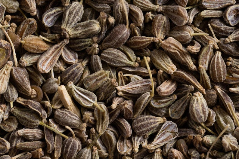 K&ouml;nnte beinhalten: Eine Ansammlung getrockneter Anissamen, die jeweils eine ausgepr&auml;gte gerippte, tropfenf&ouml;rmige Struktur mit einem br&auml;unlich-grauen Farbton aufweisen. Die Samen liegen dicht beieinander und zeigen ihre nat&uuml;rlichen, texturierten Oberfl&auml;chen.