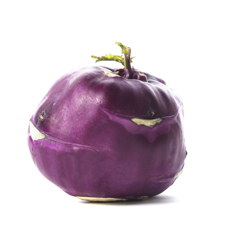 Op de afbeelding: Een enkele paarse koolrabi-knol met een gladde, ronde schil en een kleine groene scheut die uit de bovenkant steekt. De groente heeft een dieppaarse tint met subtiele lichtere vlekken en een stevige, bolvormige structuur tegen een witte achtergrond.