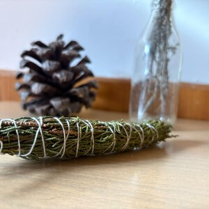 May include: A bundle of dried green cedar needles tightly wrapped with thin white cotton string. The cylindrical bundle is positioned horizontally in the foreground, with a brown pinecone and a clear glass bottle visible in the background.