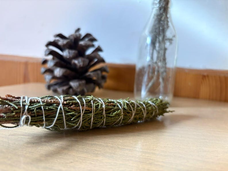 May include: A bundle of dried green cedar needles tightly wrapped with thin white cotton string. The cylindrical bundle is positioned horizontally in the foreground, with a brown pinecone and a clear glass bottle visible in the background.