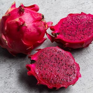 May include: One whole dragon fruit and two halves of a dragon fruit with vibrant magenta skin and deep red flesh speckled with tiny black seeds. The fruit features prominent, scale-like green-tipped leaves on the exterior of the whole specimen.