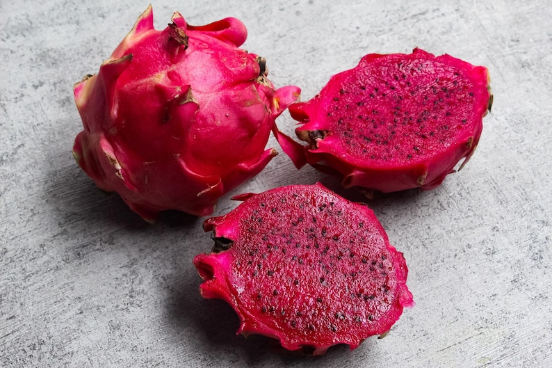 May include: One whole dragon fruit and two halves of a dragon fruit with vibrant magenta skin and deep red flesh speckled with tiny black seeds. The fruit features prominent, scale-like green-tipped leaves on the exterior of the whole specimen.