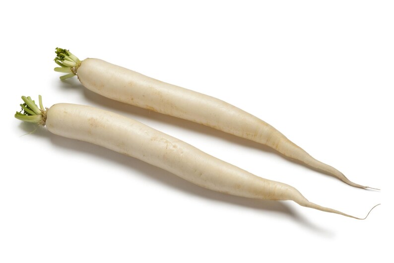 Op de afbeelding: Twee lange, cilindervormige witte daikon-radijzen met een gladde, bleke schil en kleine, lichtgroene blaadjes aan de stengeluiteinden. De groenten liggen diagonaal en lopen uit in dunne, puntige wortels aan de onderkant op een witte achtergrond.