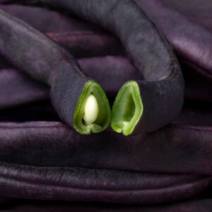 May include: A pile of deep purple beans with a smooth, elongated shape. One bean is broken open in the centre, revealing a light green interior and a single, small, pale white seed nestled inside the pod, contrasting with the dark outer skin.