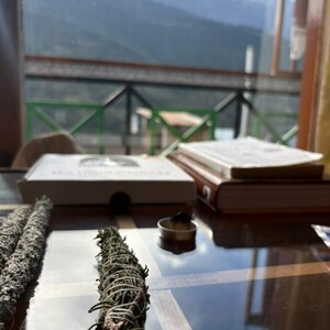 May include: Two bundles of dried green sage wrapped in white string are positioned on a reflective surface. Nearby, a small metal container holds a burning incense cone. In the background, a white box and a thick book rest on the table.