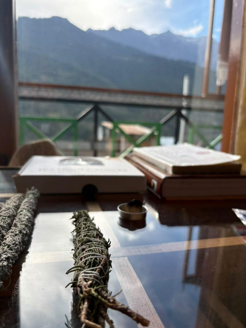 May include: Two bundles of dried green sage wrapped in white string are positioned on a reflective surface. Nearby, a small metal container holds a burning incense cone. In the background, a white box and a thick book rest on the table.