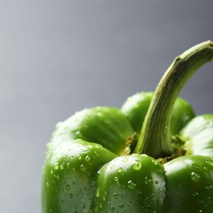Puede incluir: Un pimiento verde fresco con una piel lisa y brillante cubierta de peque&ntilde;as gotas de agua. La verdura presenta un tallo verde prominente y robusto en la parte superior, con profundas crestas verticales y l&oacute;bulos redondeados visibles.