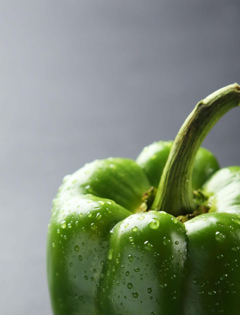 Puede incluir: Un pimiento verde fresco con una piel lisa y brillante cubierta de peque&ntilde;as gotas de agua. La verdura presenta un tallo verde prominente y robusto en la parte superior, con profundas crestas verticales y l&oacute;bulos redondeados visibles.