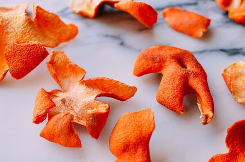 May include: A collection of dried orange citrus peels scattered across the frame. Each piece of peel displays a vibrant, textured orange exterior with a porous surface and a lighter, pale white pith on the underside, showing irregular shapes.