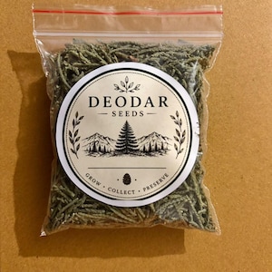 May include: A clear plastic resealable bag filled with dried green Deodar cedar needles. A circular white label on the front features black text reading DEODAR SEEDS, GROW COLLECT PRESERVE, and a graphic illustration of a mountain range and pine tree.