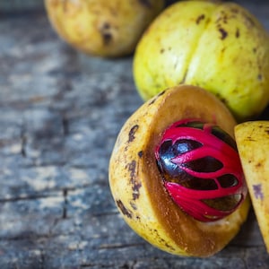 Puede incluir: Varios frutos de nuez moscada con c&aacute;scaras exteriores amarillas y texturizadas. Un fruto est&aacute; abierto para revelar una semilla de color marr&oacute;n oscuro parcialmente envuelta en un arilo rojo brillante, que contrasta con el interior amarillo p&aacute;lido.