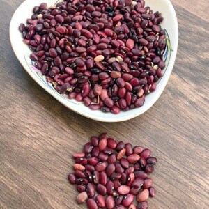 May include: A white, teardrop-shaped bowl filled with dried dark red kidney beans, accompanied by a small, loose pile of the same kidney beans positioned directly below the bowl on the surface.