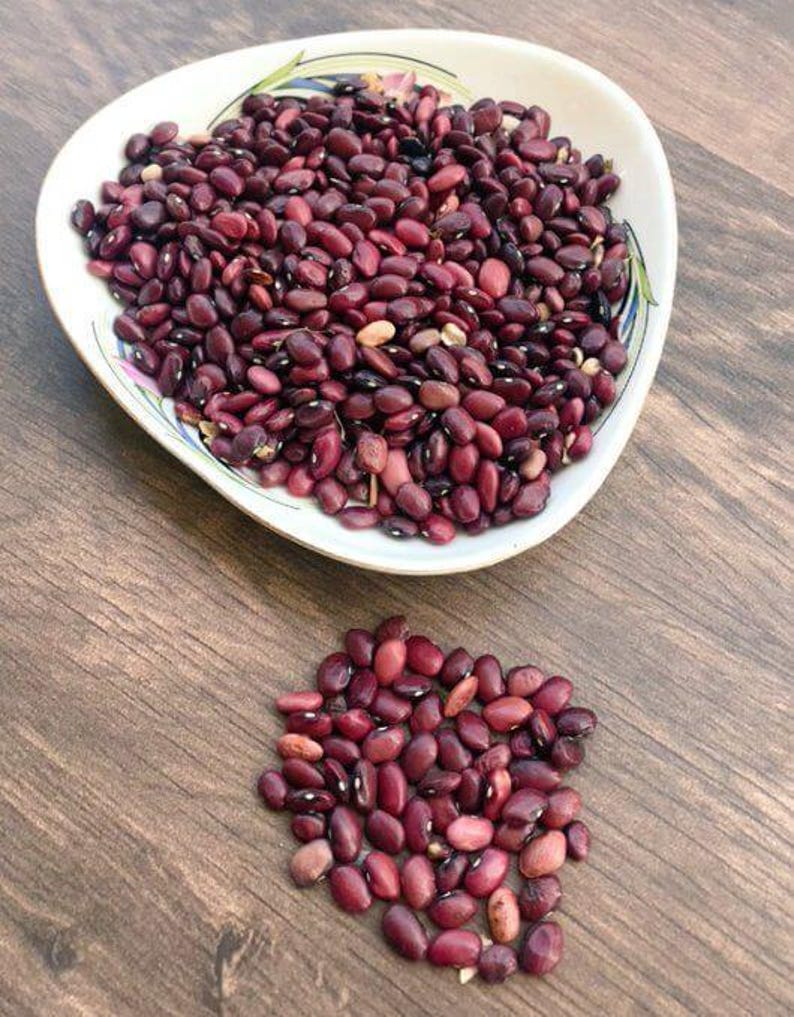 May include: A white, teardrop-shaped bowl filled with dried dark red kidney beans, accompanied by a small, loose pile of the same kidney beans positioned directly below the bowl on the surface.