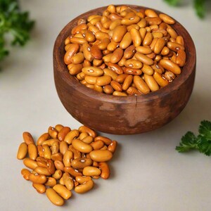 Puede incluir: Un cuenco de madera marr&oacute;n oscuro lleno de numerosos frijoles de color marr&oacute;n claro, con un peque&ntilde;o mont&oacute;n de los mismos frijoles descansando fuera del cuenco. Los frijoles son de forma ovalada con una textura suave y una marca blanca.