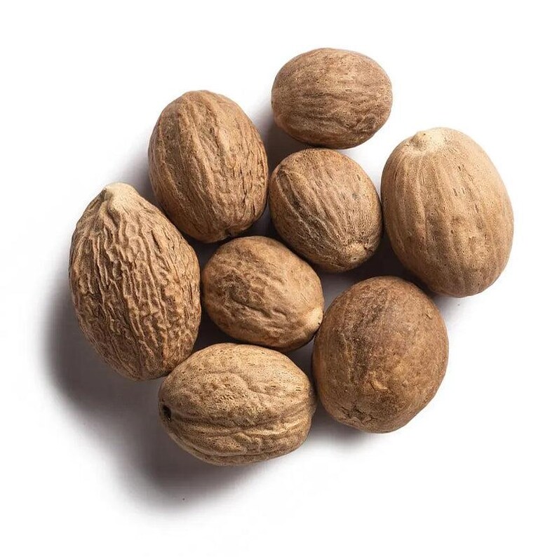 Puede incluir: Un grupo de ocho nueces moscadas enteras con un exterior de color marr&oacute;n claro, texturizado y le&ntilde;oso. Cada semilla tiene forma ovalada con surcos naturales visibles y una superficie seca y mate, dispuestas en un grupo sobre fondo blanco.