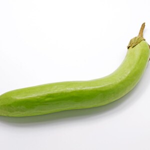 Puede incluir: Una berenjena larga y delgada de color verde claro con un cuerpo liso ligeramente curvado y un peque&ntilde;o tallo marr&oacute;n texturizado en la parte superior. La verdura tiene un tono de piel verde lima p&aacute;lido uniforme y un extremo redondeado.