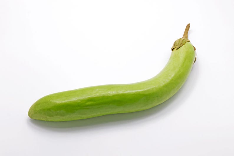 Puede incluir: Una berenjena larga y delgada de color verde claro con un cuerpo liso ligeramente curvado y un peque&ntilde;o tallo marr&oacute;n texturizado en la parte superior. La verdura tiene un tono de piel verde lima p&aacute;lido uniforme y un extremo redondeado.