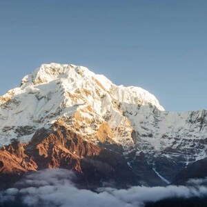 May include: A snow-capped mountain range featuring a prominent central peak with bright white glaciers and rugged, dark reddish-brown rock formations at the base, partially obscured by low-hanging white clouds against a clear blue sky.