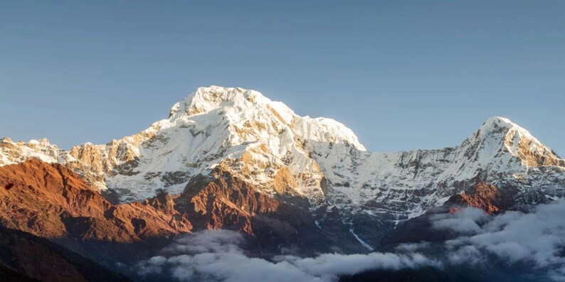 May include: A snow-capped mountain range featuring a prominent central peak with bright white glaciers and rugged, dark reddish-brown rock formations at the base, partially obscured by low-hanging white clouds against a clear blue sky.